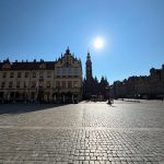 Słoneczny, szeroki widok na Rynek we Wrocławiu, pokazujący wieżę Starego Ratusza, zabytkowe kamienice i bruk pod czystym błękitnym niebem.