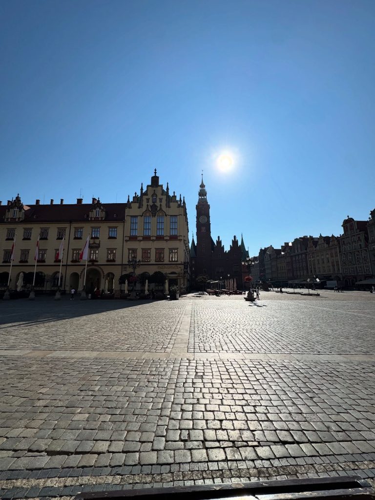 Słoneczny, szeroki widok na Rynek we Wrocławiu, pokazujący wieżę Starego Ratusza, zabytkowe kamienice i bruk pod czystym błękitnym niebem.